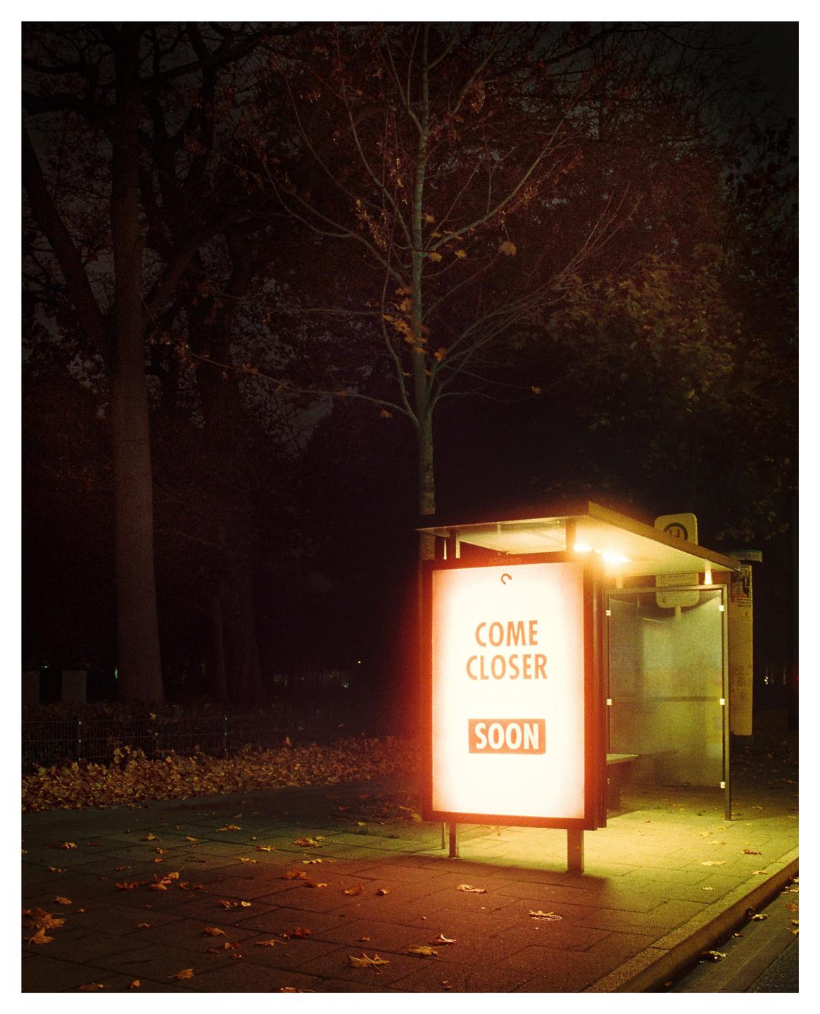 A bus stop with an illuminated advertisement sign that reads "COME CLOSER SOON" is visible at night.