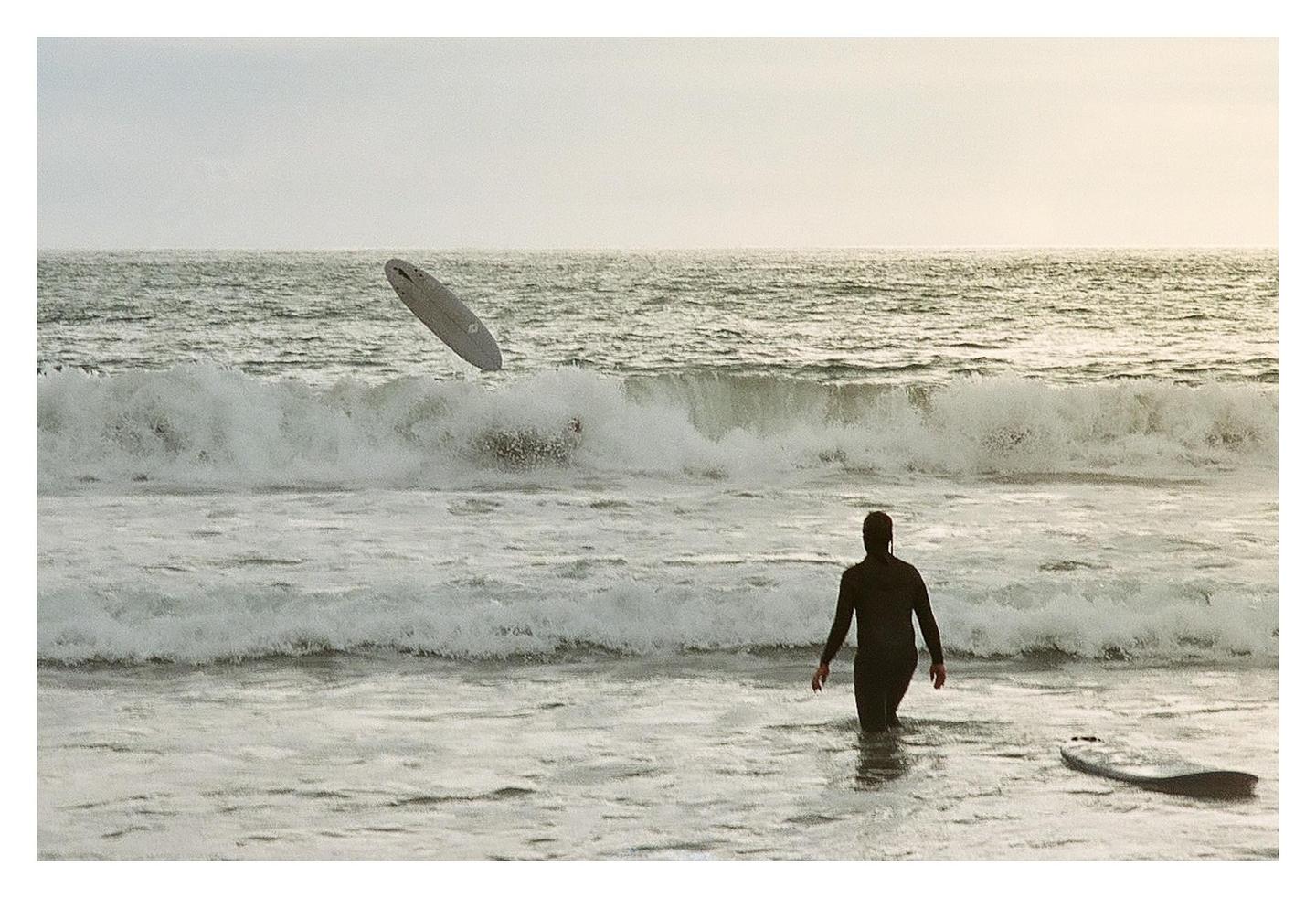 A surfer in a wetsuit walks into the ocean as a surfboard flies through the air above a breaking wave.