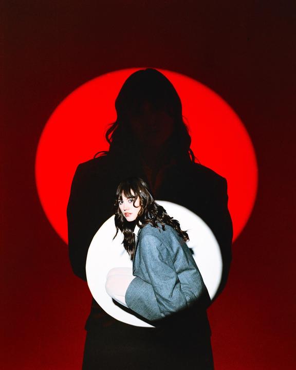 A woman crouches in a white spotlight, casting a large shadow against a red spotlight on the wall behind her.