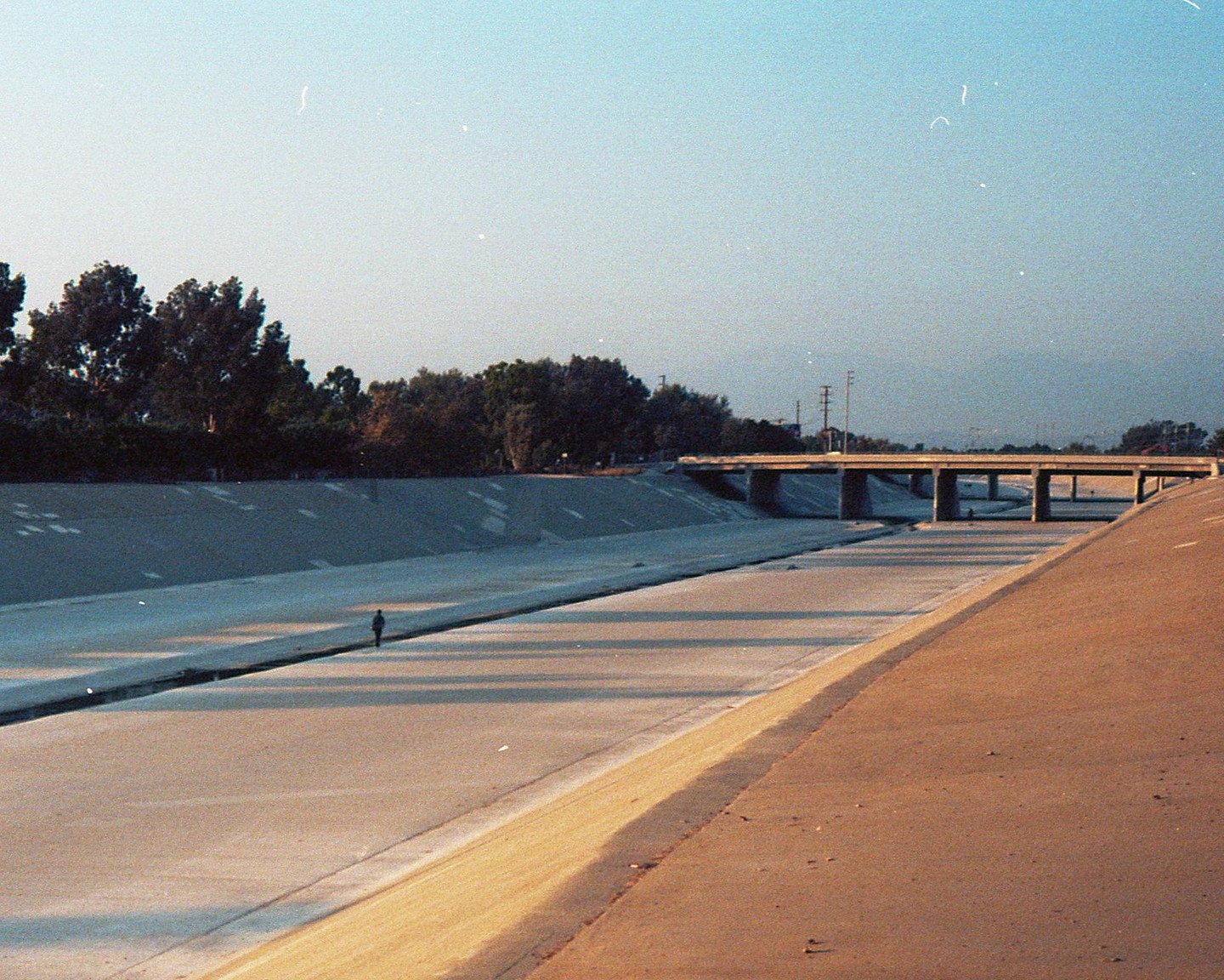A person walks along a concrete riverbed under a bridge with trees lining the banks.