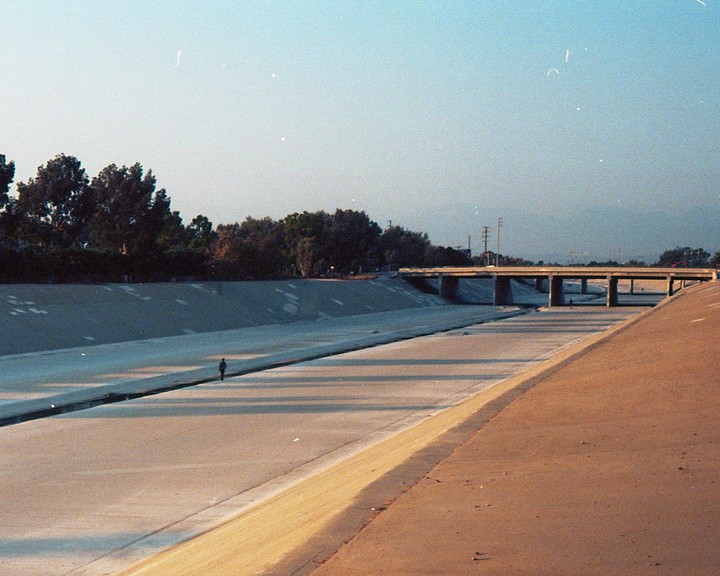 A person walks along a concrete riverbed under a bridge with trees lining the banks.