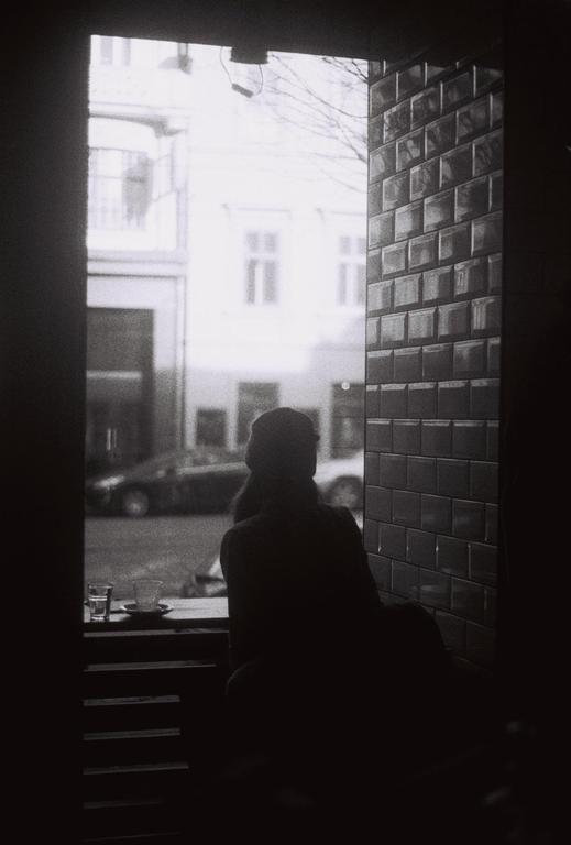 A person sits in a dark room looking out a window at a street with buildings and a car.