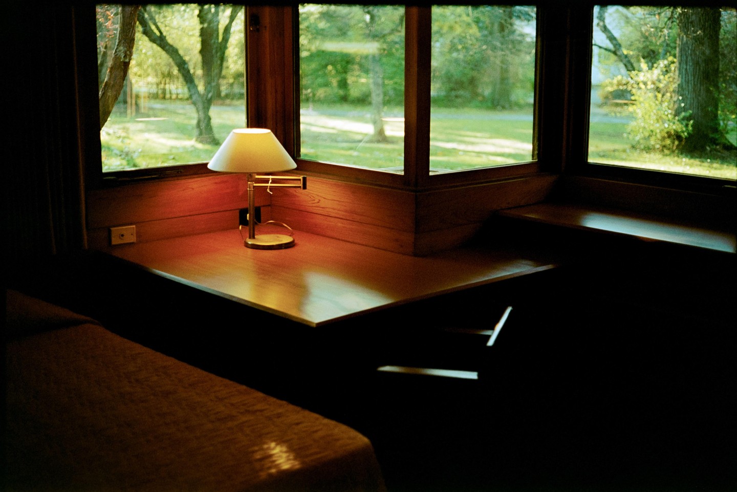 A lamp sits on a wooden desk next to a window looking out onto a green lawn and trees.