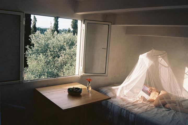 A woman reads a book in bed under a mosquito net next to a window overlooking green trees.
