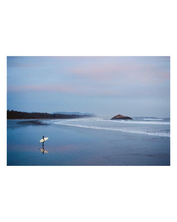 A surfer stands on a beach with a surfboard, reflected in the wet sand, with an island and forest in the distance under a pink and blue sky.