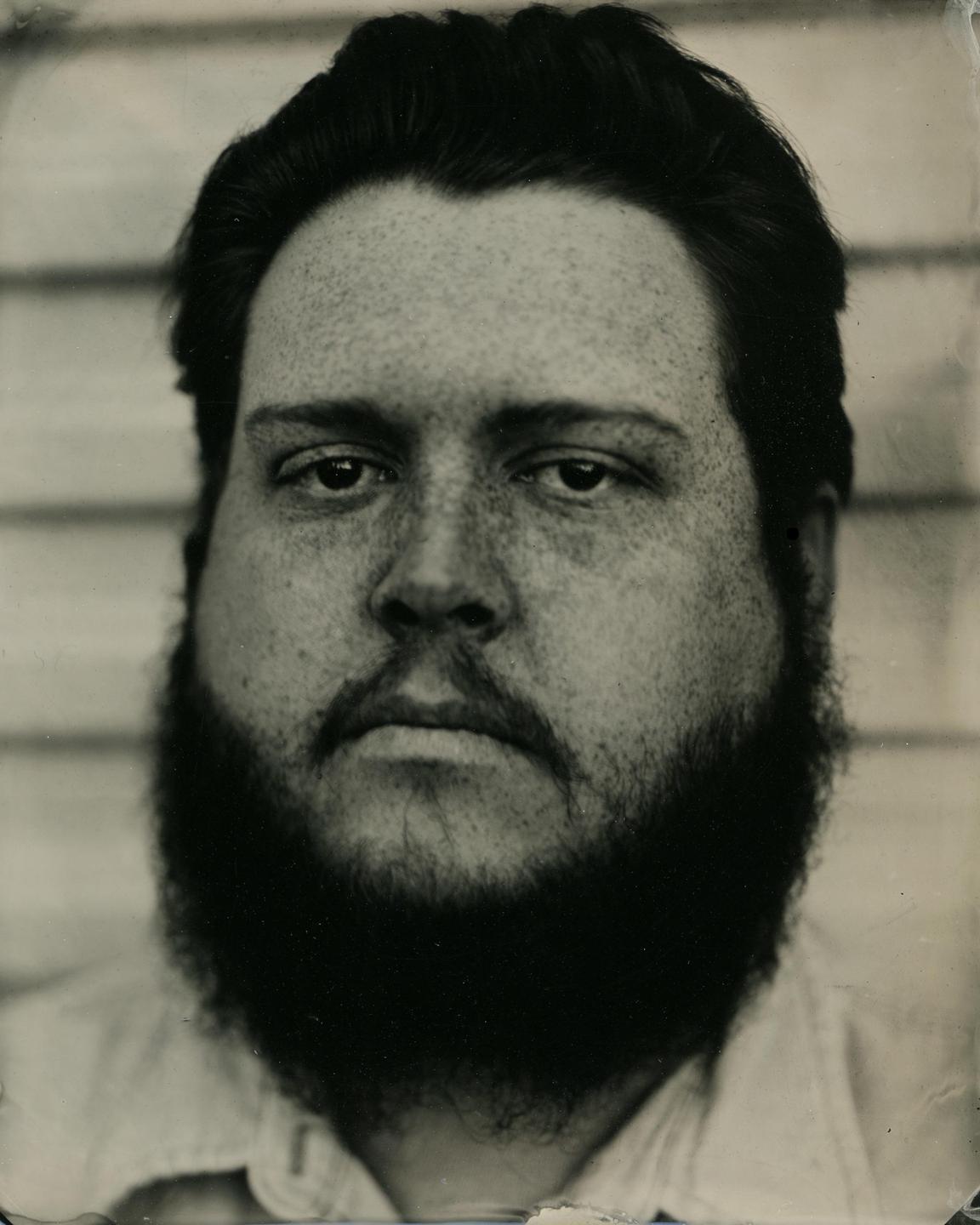A man with a beard and freckles looks directly at the viewer in a close-up portrait.