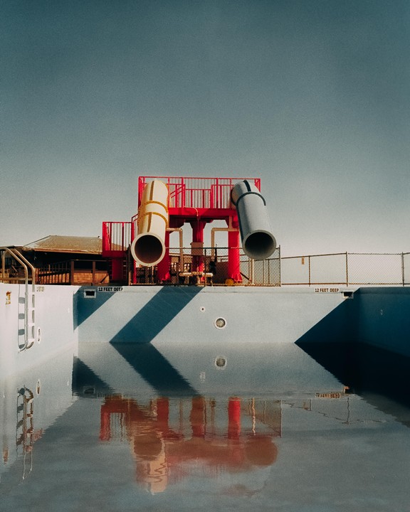 A red and yellow water slide stands over an empty swimming pool with a reflection of the slide in the water.
