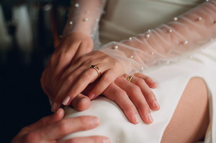 A couple's hands are intertwined, with the woman wearing a sheer, pearl-embellished sleeve and a ring with a dark stone, and the man wearing a gold band.