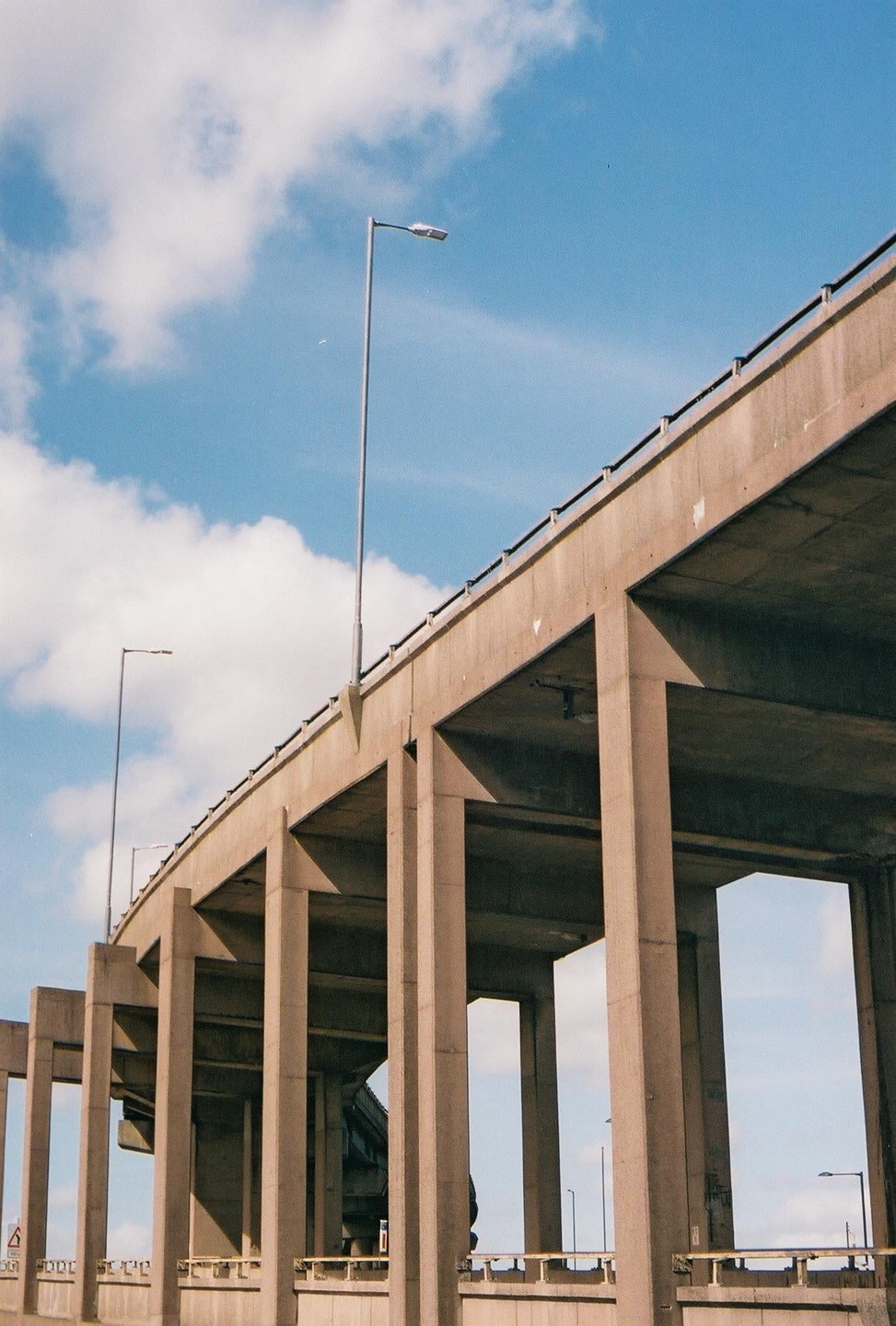 A concrete highway overpass with streetlights is viewed from below against a blue sky with white clouds.