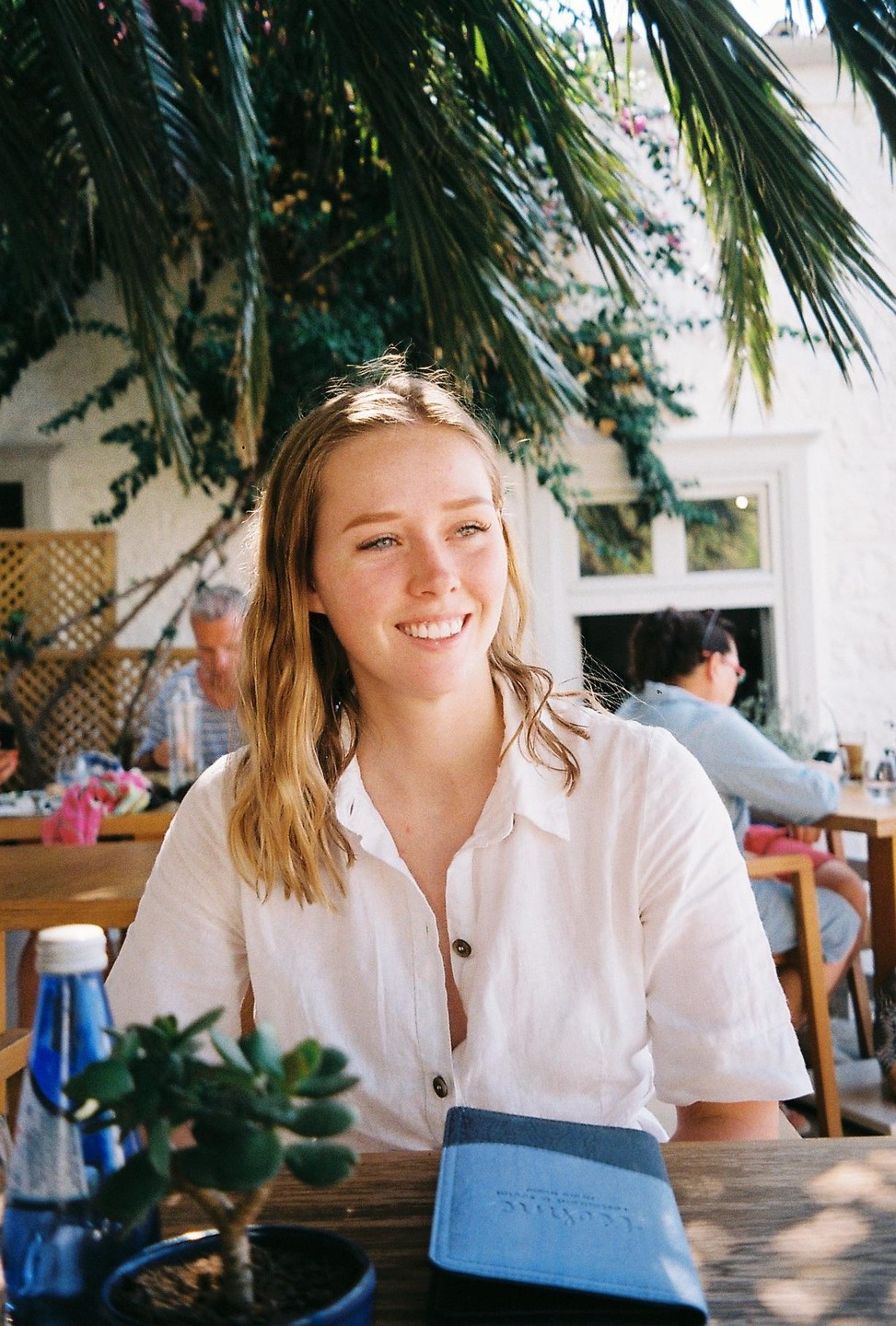 A young woman with long hair smiles while sitting at a wooden table with a small plant and a blue bottle.