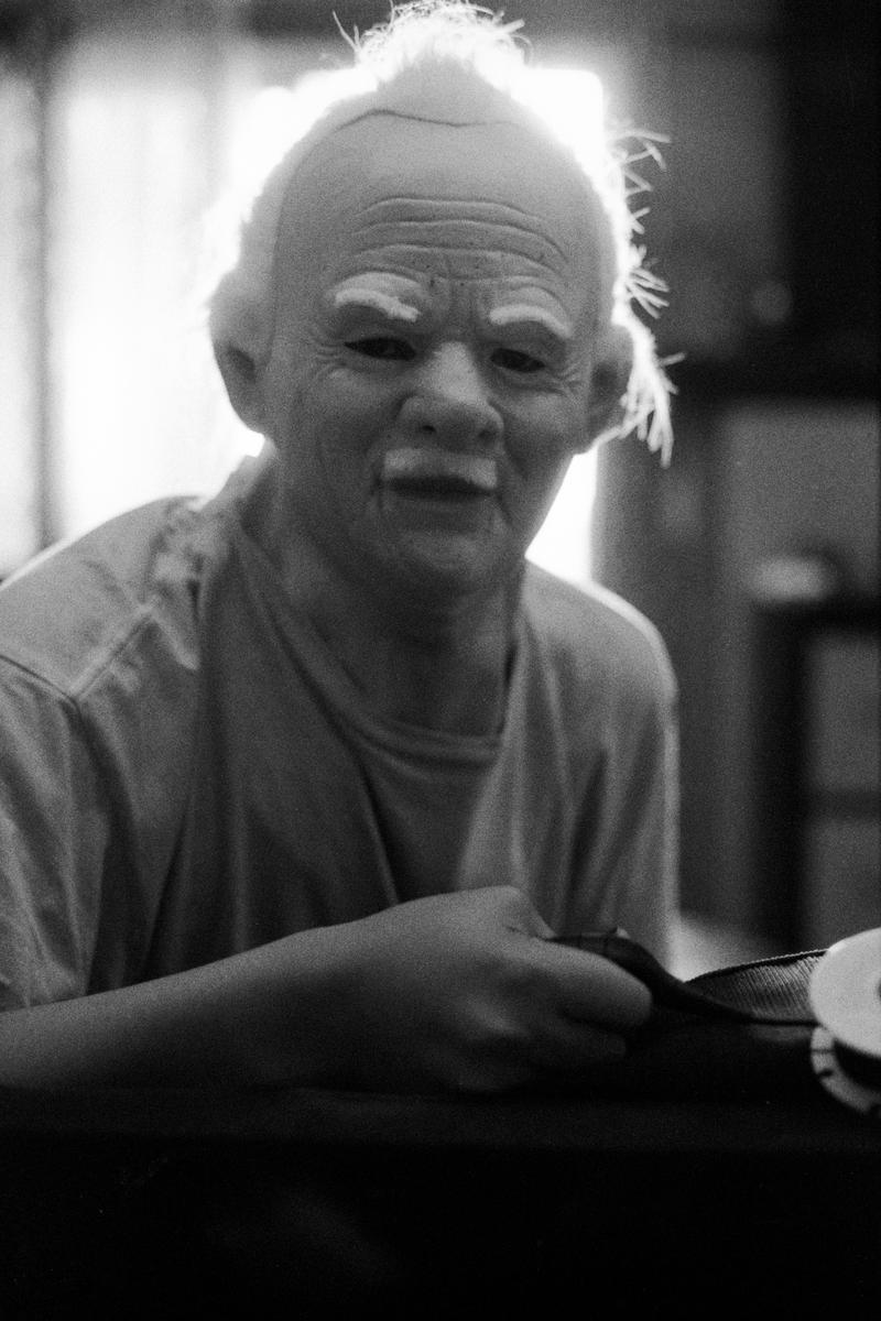 A person wearing an old man mask is backlit, holding a ribbon.