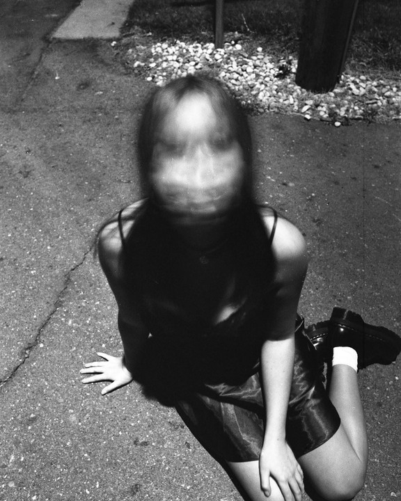 A woman with a blurred face sits on asphalt next to a patch of gravel and a dark pole.