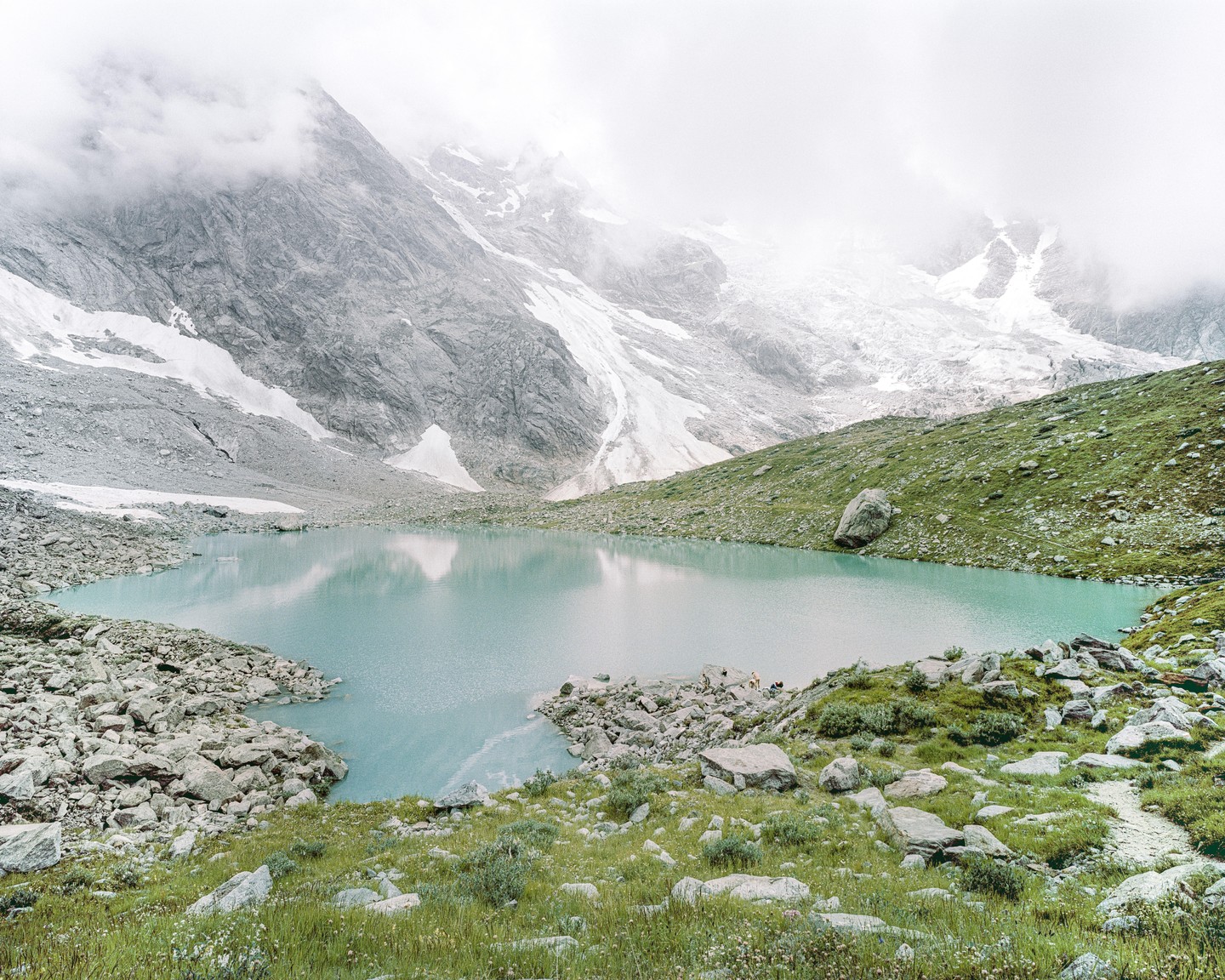 A glacial lake is surrounded by rocky and grassy terrain with snow-capped mountains in the background.