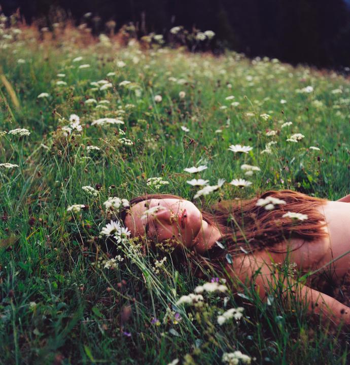 A woman with long hair lies in a field of tall grass and wildflowers.