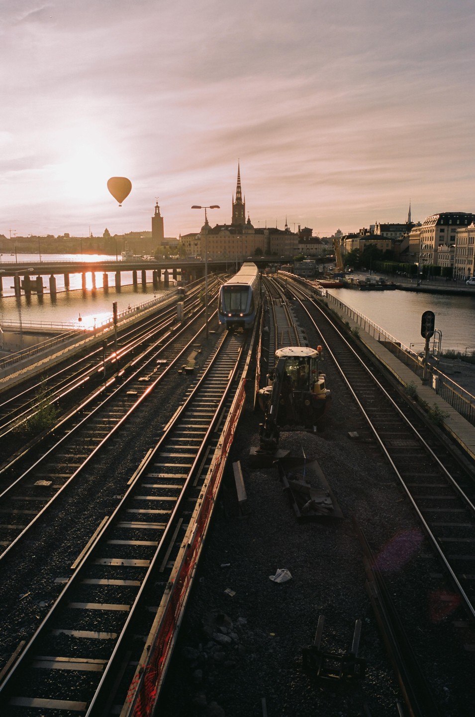 A hot air balloon floats over a city skyline with a train on tracks in the foreground at sunset.