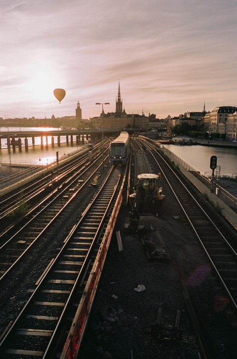 A hot air balloon floats over a city skyline with a train on tracks in the foreground at sunset.