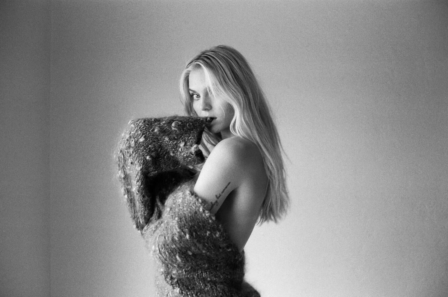 A woman with long hair is wrapped in a textured sweater, looking over her shoulder.
