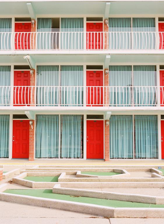 A three-story motel with red doors and light blue curtains has a miniature golf course in front of it.