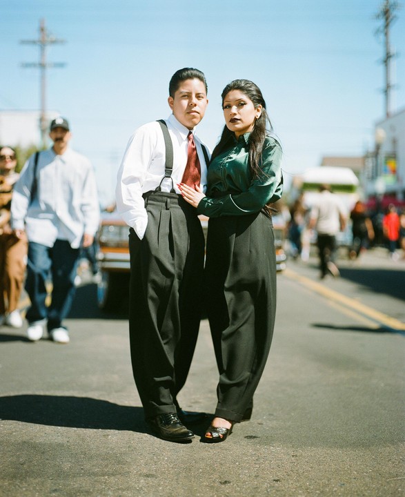A couple dressed in vintage-style clothing stands on a street with other people in the background.