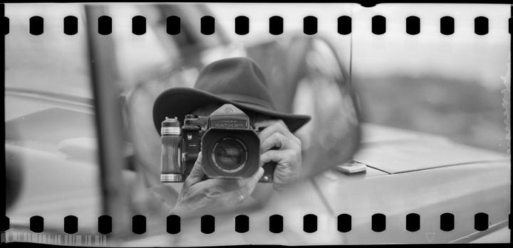A person wearing a hat holds a camera with a reflection of the camera in the lens.