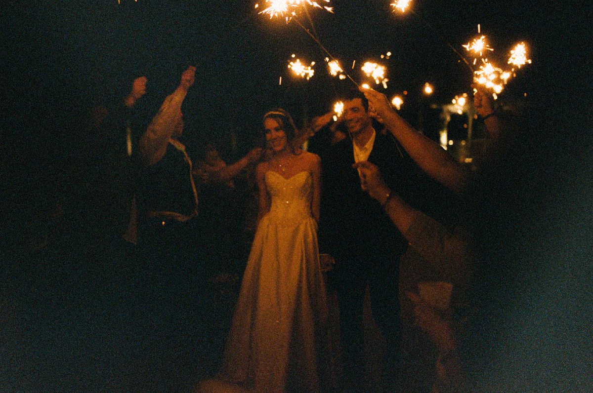 A couple walks through a crowd of people holding sparklers at night.