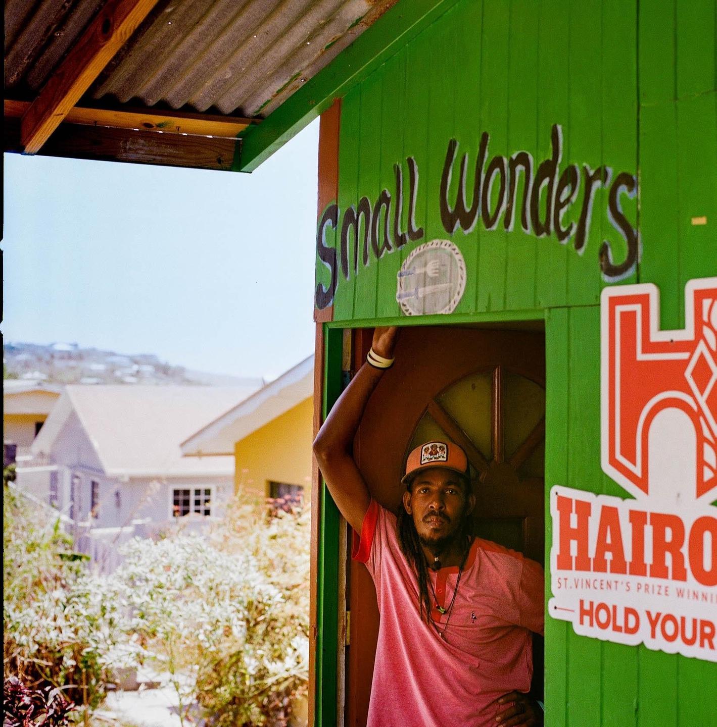 A man stands in the doorway of a green building with "Small Wonders" written on it.