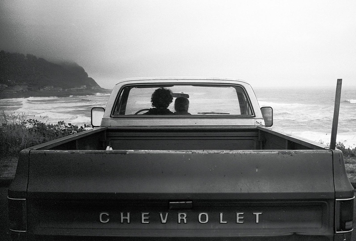 Two people sit in a Chevrolet pickup truck overlooking a wavy ocean and a distant coastline.