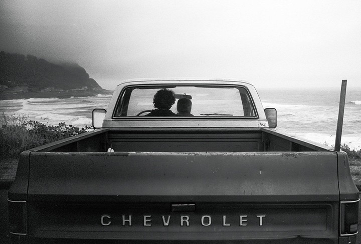 Two people sit in a Chevrolet pickup truck overlooking a wavy ocean and a distant coastline.
