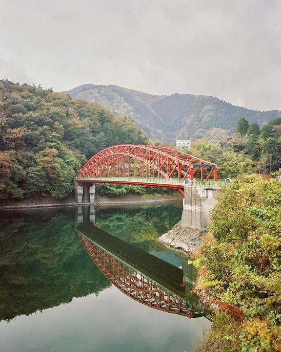 A red arch bridge spans a body of water, reflecting in the still surface, with forested mountains in the background.