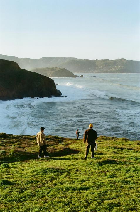 Three men stand on a grassy cliff overlooking a wavy ocean with distant hills under a clear sky.