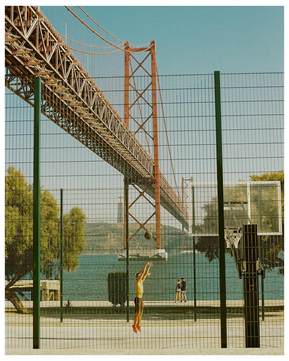 A man jumps to shoot a basketball on an outdoor court next to a body of water with a large bridge overhead.