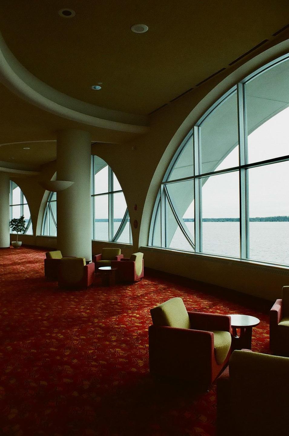 A large room with a red patterned carpet features several armchairs and tables, with arched windows overlooking a body of water.