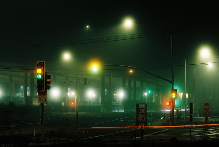 A foggy night scene shows a street with traffic lights and a highway overpass in the background.