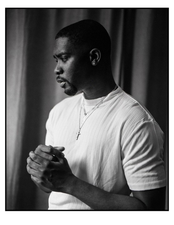 A man with clasped hands looks down and to the left, wearing a white shirt and a cross necklace.
