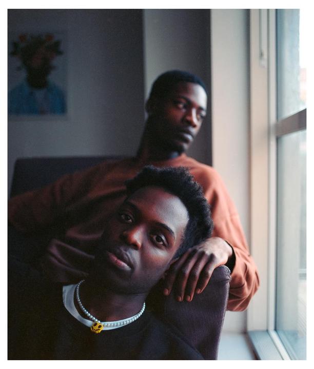A man with a pearl necklace rests his head on a chair while another man sits behind him looking out a window.