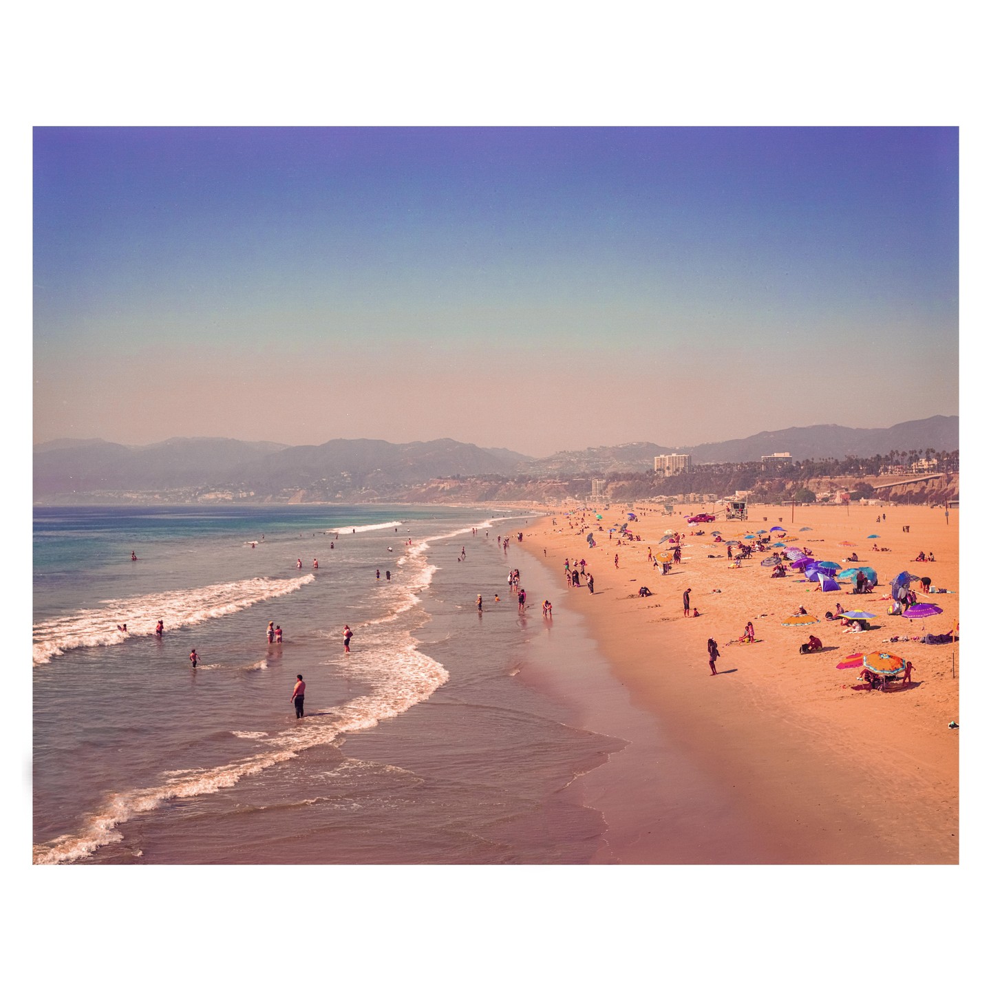 A wide view shows many people on a beach with waves rolling in and mountains in the distance.
