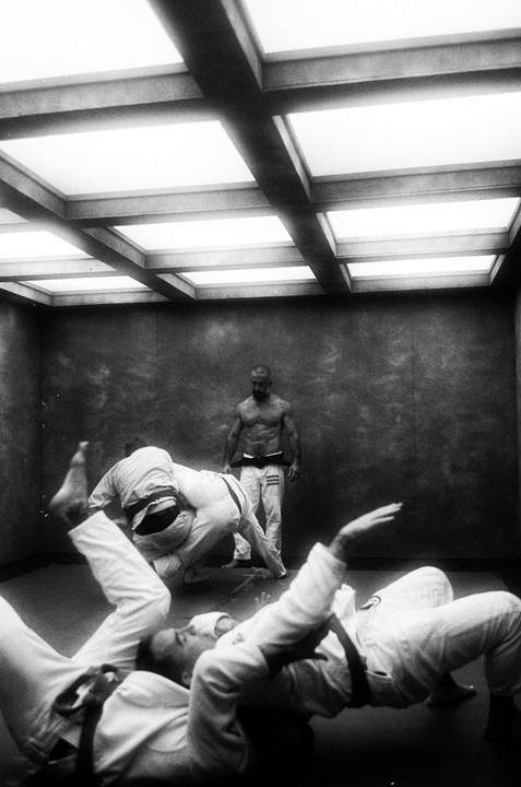 Three men are practicing jiu-jitsu in a room with a grid of bright ceiling lights.
