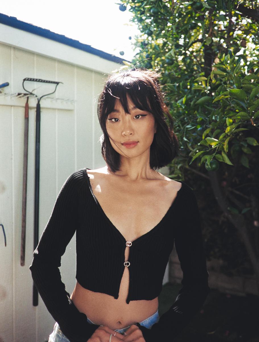 A woman with dark hair and a black top stands in a garden with a white shed and green trees in the background.