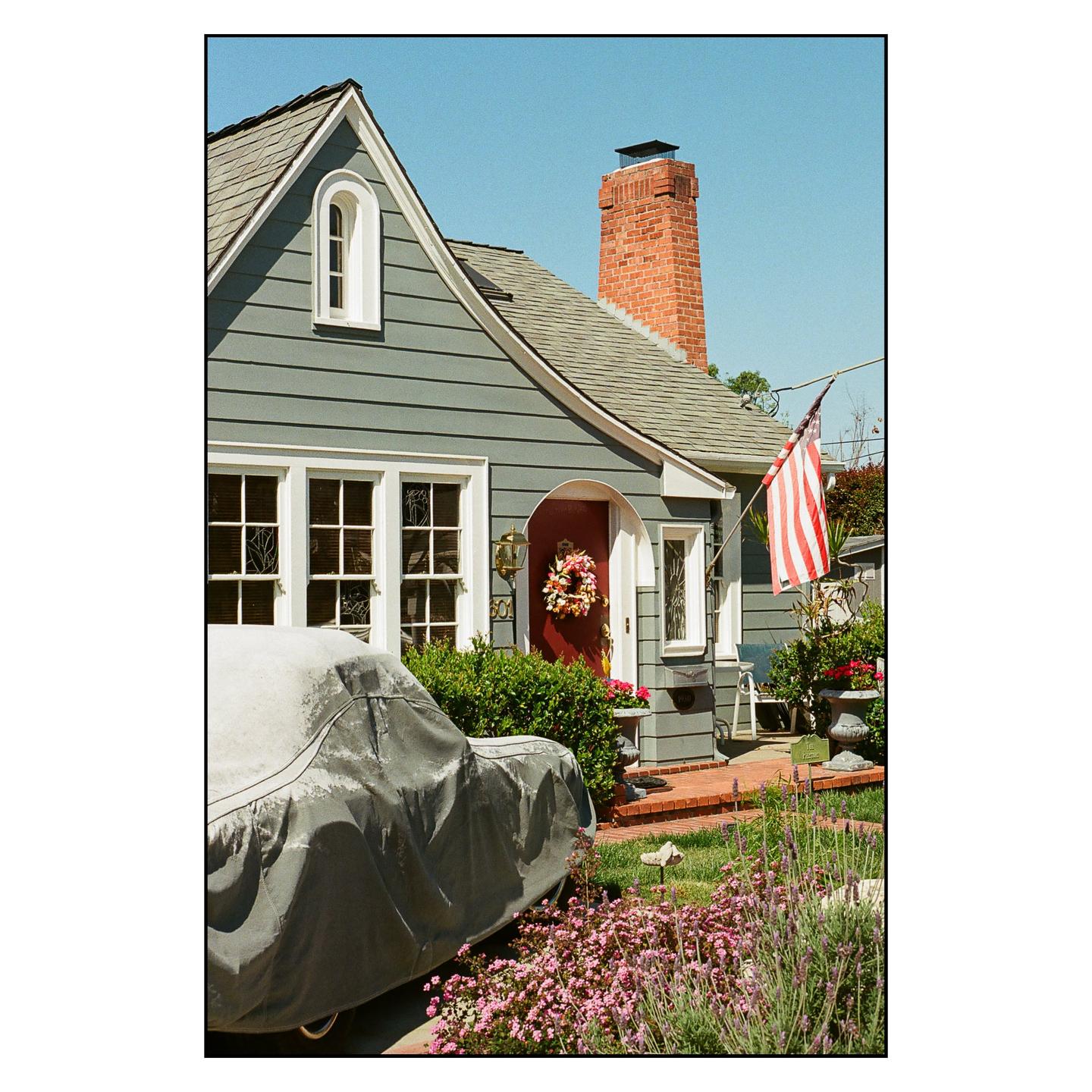 A house with a red door and an American flag has a car covered with a tarp parked in front of it.