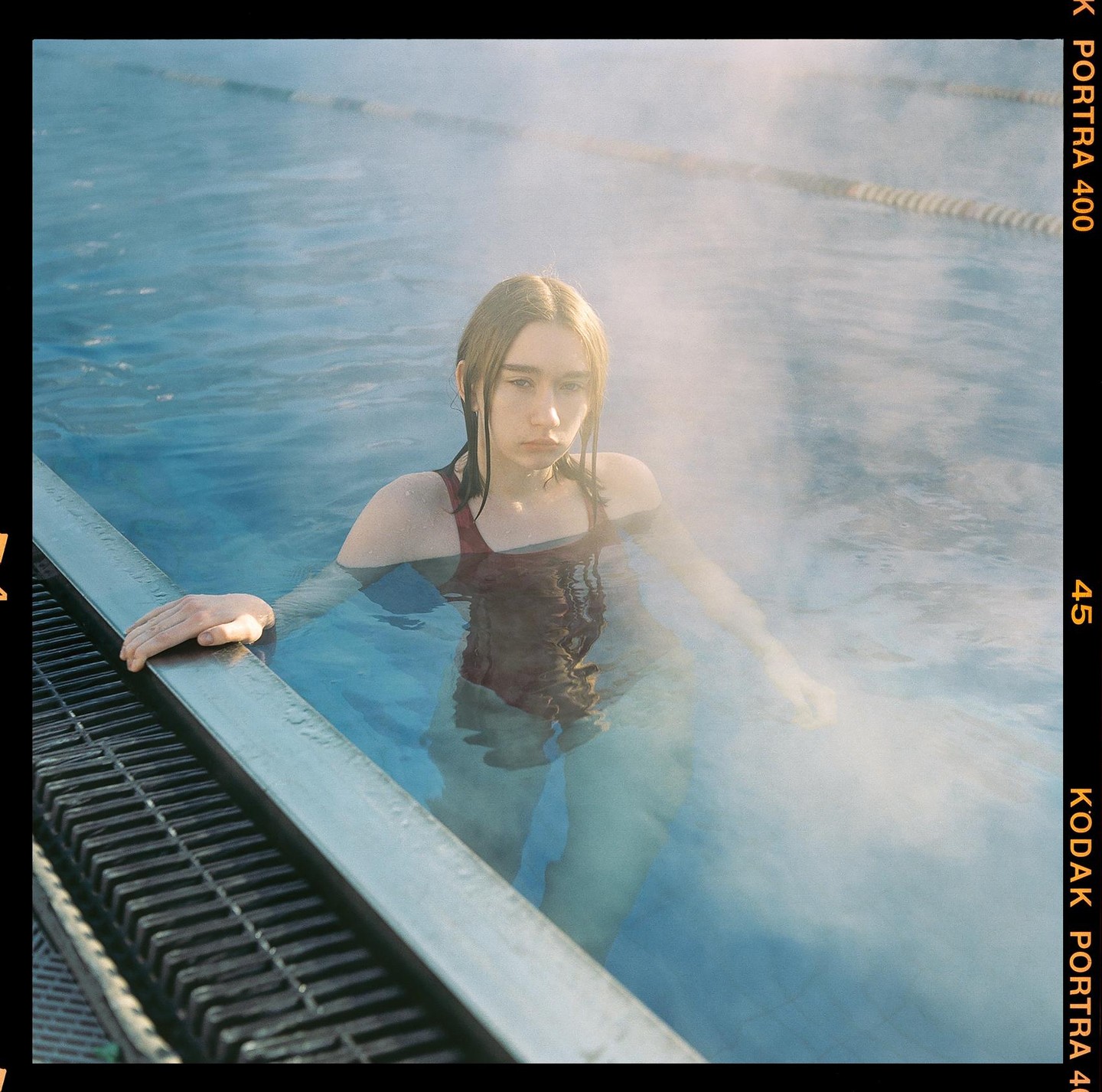 A young woman in a red swimsuit stands in a steaming outdoor swimming pool.