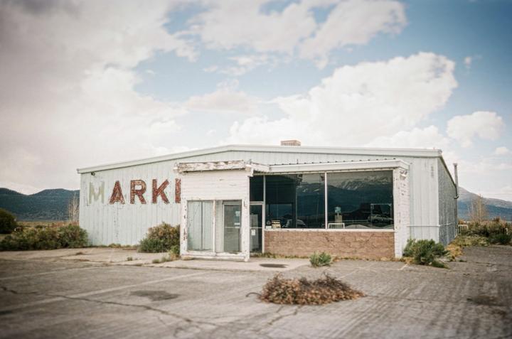 An abandoned building with "MARKI" painted on its side stands under a cloudy sky, with mountains visible in the distance.