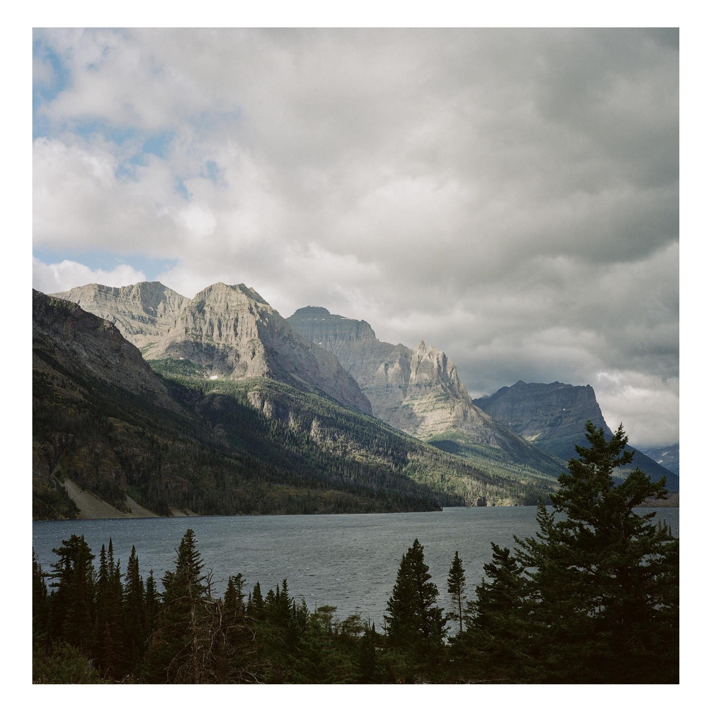 A lake is surrounded by a forest and mountains under a cloudy sky.