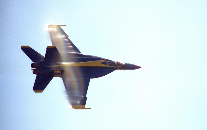 A jet aircraft with blue and yellow markings flies through a bright sky with a rainbow lens flare.