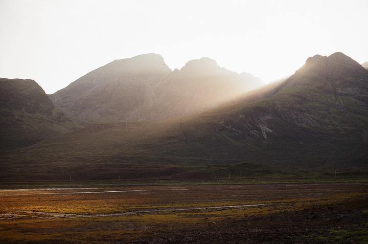 Sunlight streams over a mountain range and illuminates a field in the foreground.