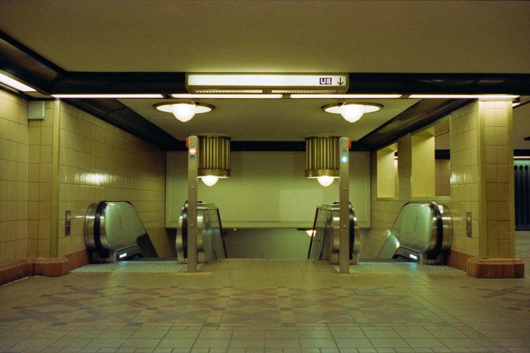 Two escalators descend into a subway station with tiled walls and patterned floor.