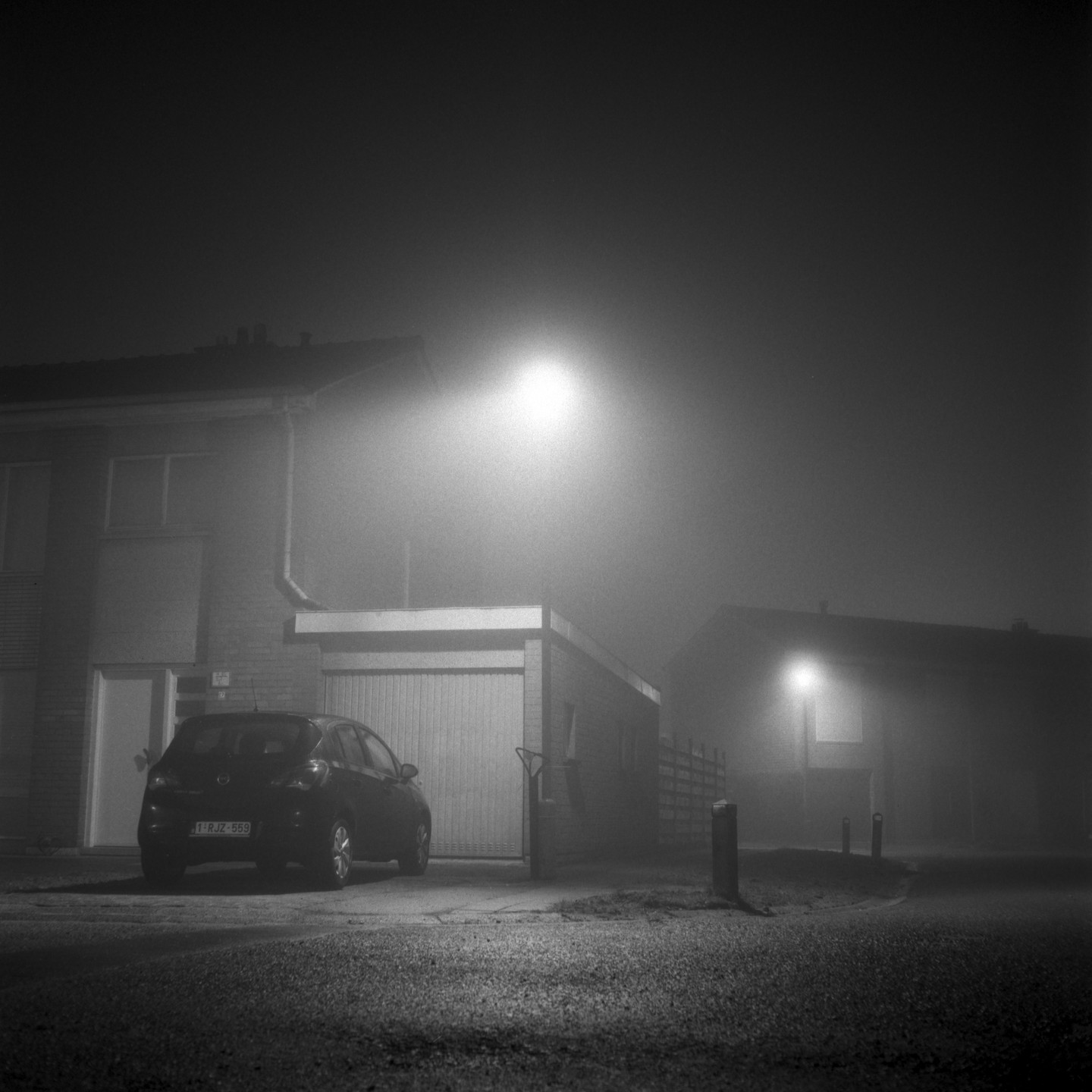 A car is parked in front of a garage and house on a foggy night under a streetlamp.