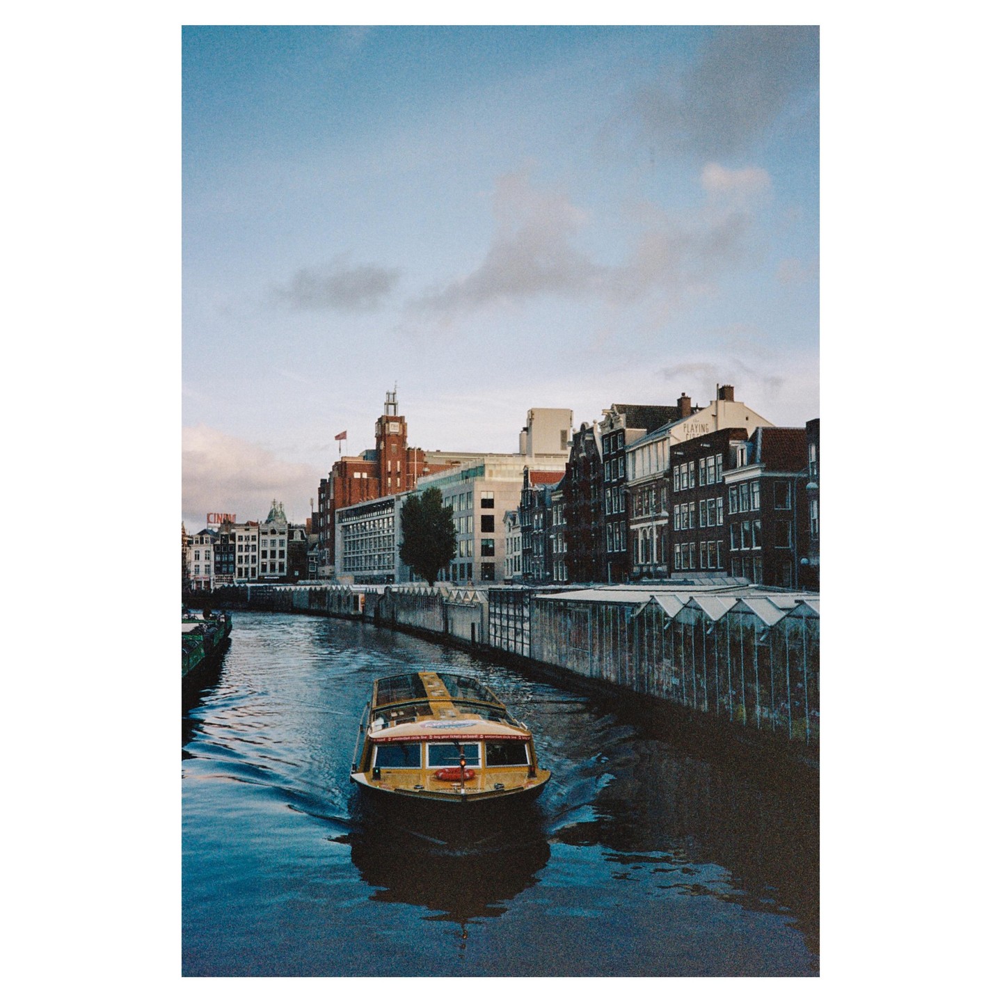 A yellow boat travels down a canal lined with buildings and greenhouses under a cloudy sky.