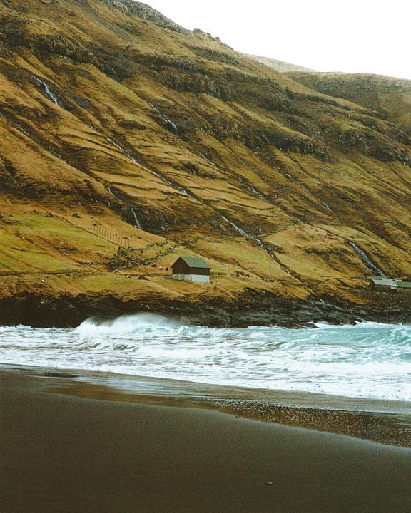 A small house with a green roof sits on a grassy hillside overlooking a black sand beach with crashing waves.