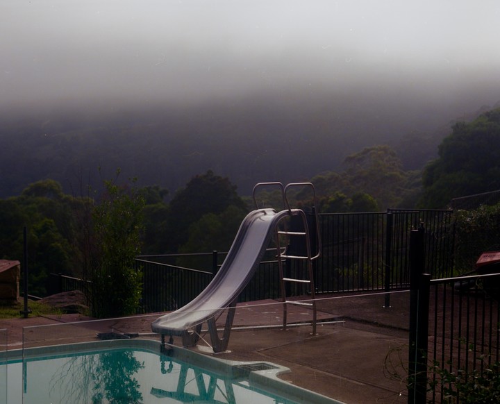 A swimming pool with a slide overlooks a valley filled with trees and fog.