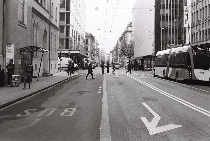 A street scene shows people crossing a road with a bus on the right and buildings lining both sides.
